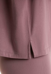 Haut mauve à manches courtes en tissu doux, présentant une fente sur le côté à l'ourlet et des coutures nettes le long des bords.
