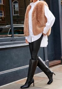 Un gilet en fausse fourrure beige porté par-dessus une chemise blanche à boutons, associé à un legging en cuir noir et de hauts bottes noires à talons, avec des bouts pointus.