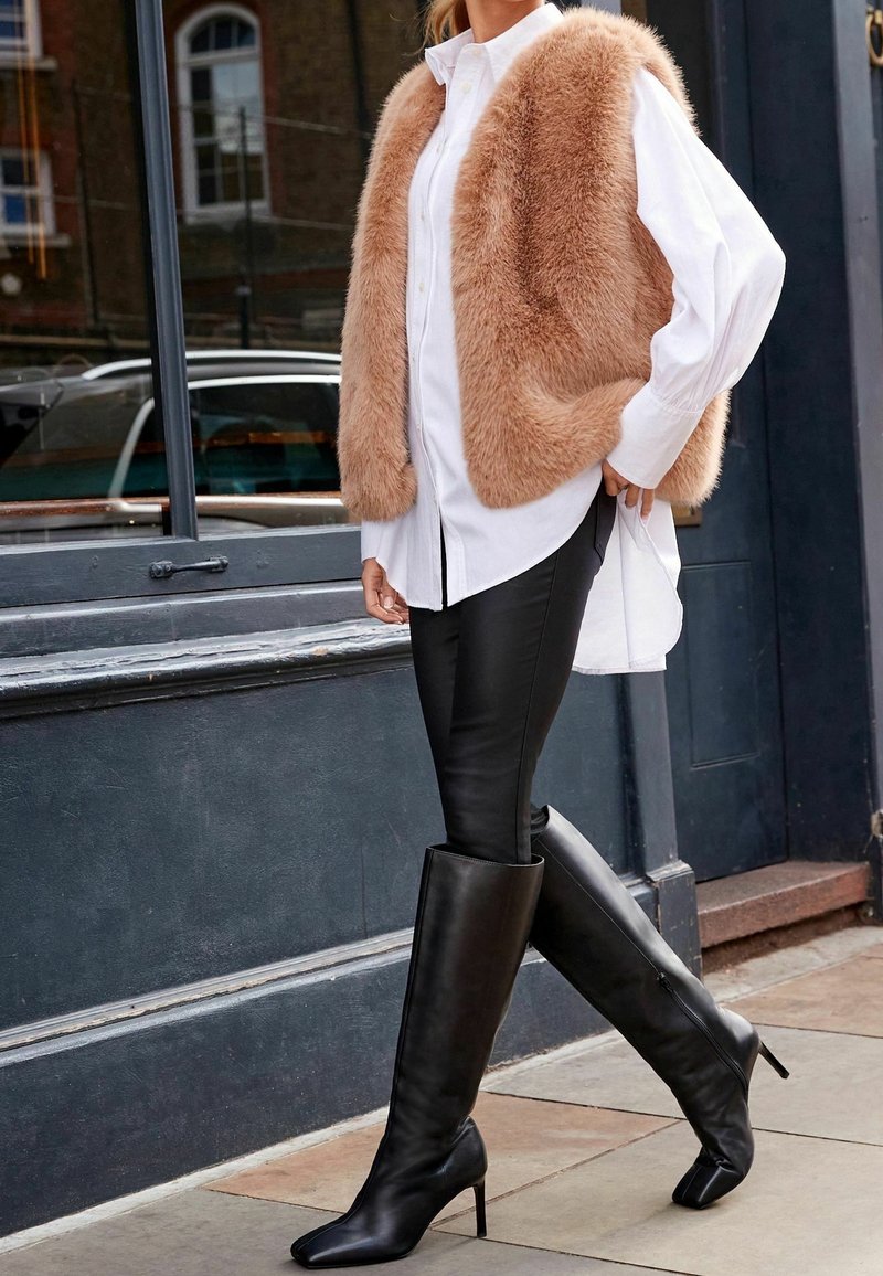 Un gilet en fausse fourrure beige porté par-dessus une chemise blanche à boutons, associé à un legging en cuir noir et de hauts bottes noires à talons, avec des bouts pointus.