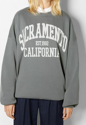 Personne portant un sweat-shirt gris avec "SACRAMENTO EST. 1982 CALIFORNIA" en lettres blanches, assorti à un pantalon foncé à fines rayures.