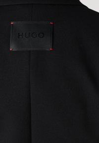 Crna sakoić s teksturiranim završetkom. Sadrži crnu kožnu oznaku s ugraviranim "HUGO", naglašenu crvenim šavovima.