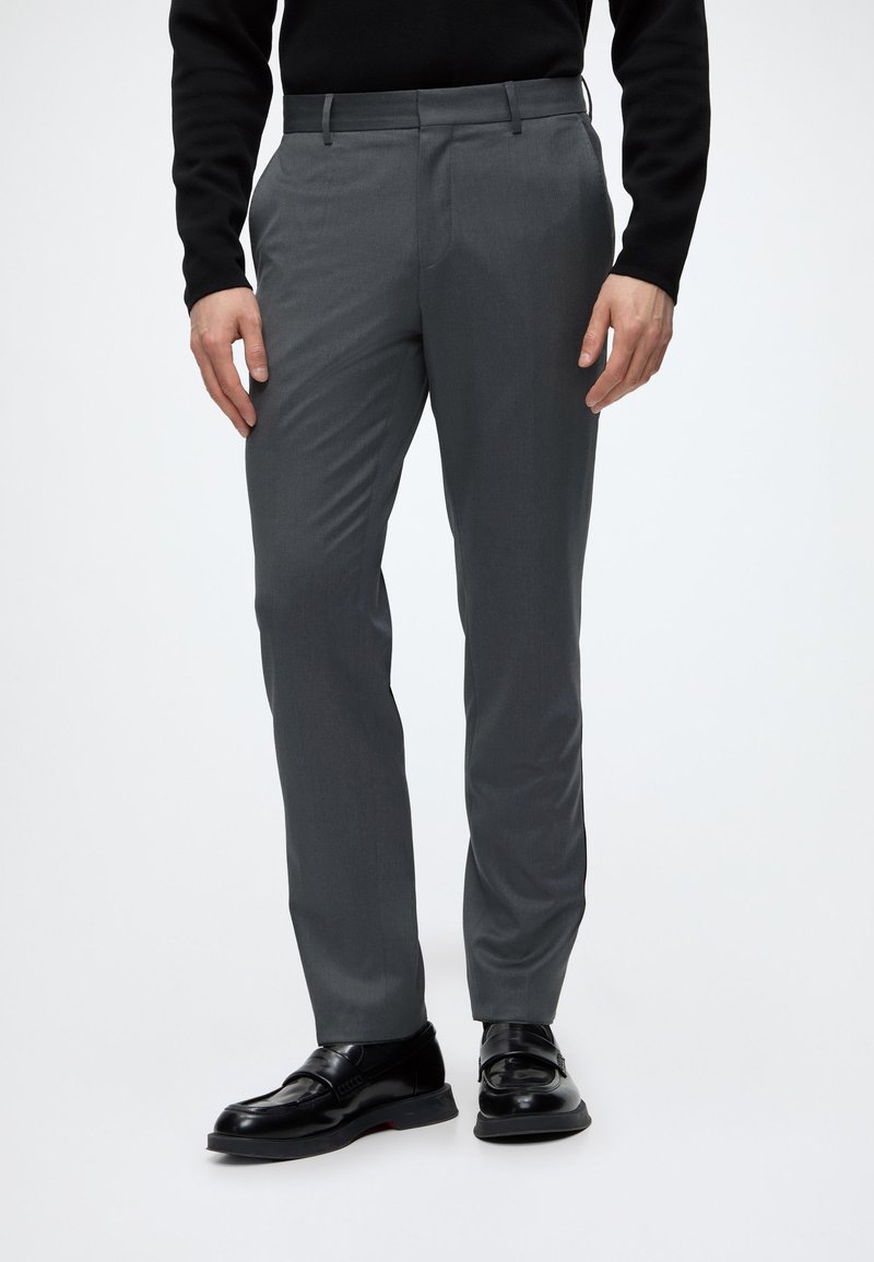 Homme portant un pantalon gris droit, une chemise noire à manches longues et des chaussures de ville en cuir noir, debout devant un fond uni.