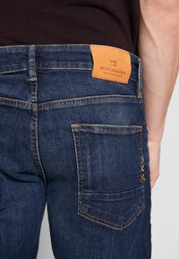 Jeans de mezclilla azul oscuro con costuras amarillas, una etiqueta de cuero en la parte trasera de la cinturilla y un bolsillo trasero con un patrón texturizado.