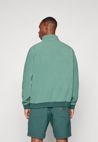 Grønn teksturert genser med høy krage, ribbestrikket kant og logo-detalj bak, sammen med matchende grønne shorts med lappelommer.