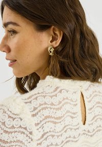 Pendientes dorados con un diseño texturizado entrelazado. El fondo muestra una prenda de encaje blanca con patrones en forma de ondas y círculos.