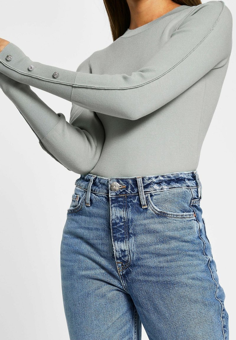 Light green long-sleeve top with ribbed texture and decorative buttons at the cuffs, paired with high-waisted blue distressed jeans.
