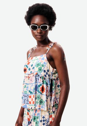 Femme portant un top fleuri patchwork sans manches et un bas assorti avec des bretelles nouées aux épaules et des lunettes de soleil ovales blanches, regardant légèrement vers la droite.