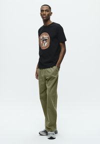 Schwarzes T-Shirt mit einem bunten Grafikdesign, kombiniert mit lockeren olivgrünen Hosen und grauen Sportschuhen.