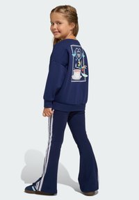 Tamnoplava dukserica s grafikom na leđima, uparena s tamnoplavim proširenim hlačama koje imaju bijele bočne pruge. Materijal izgleda mekan i ugodan.