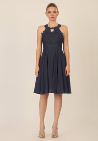 Marineblaues Kleid mit einem bodenlangen floralen Spitzenoberteil und einem Ausschnitt in Herzform; ausgestellter Rock aus leichtem Stoff; kombiniert mit silbernen Riemchen-Heels.