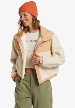 Fleece-lined jacket in cream with a tan nylon upper, featuring snap closures, elastic cuffs, and a logo patch. Worn with a graphic tee and green pants.