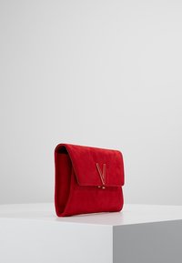 Punainen mokkainen kirjekuorilaukku, jossa on läppäkiinnitys. Edessä näkyvä kultainen "V"-koriste. Sileä pinta, minimalistinen muotoilu.