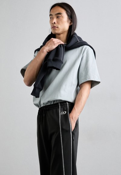 T-shirt gris clair à manches courtes associé à un pantalon de survêtement noir avec des bandes blanches sur les côtés et un logo, avec un sweat à capuche noir drapé sur les épaules.