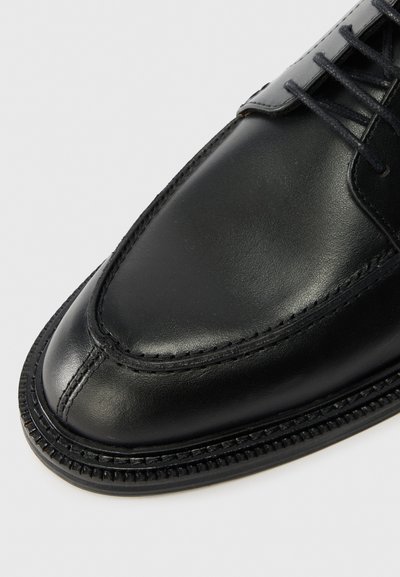 Μαύρο δερμάτινο παπούτσι, με στρογγυλή μύτη, λεία υφή και αντίθεση στη ραφή κατά μήκος της σόλας. Οι κ�ορδόνες είναι μαύρες και κομψές.