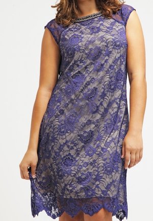 Kvinde iført ærmeløs knælang lilla blondekjole med blomsterprint og perlebesat halsudskæring, stående foran en ensfarvet baggrund.