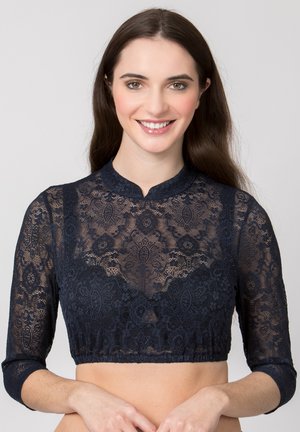 Junge Frau lächelt, trägt ein marineblaues, halbtransparentes Spitzen-Crop-Top mit dreiviertel Ärmeln und hohem Kragen.