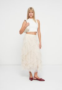 Kvinne i en hvit ermeløs crop topp og lagdelt beige tyllskjørt holder en hvit veske på skulderen, iført røde spisse sko på en hvit bakgrunn.
