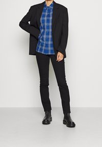 Blazer negro sobre una camisa de cuadros azul y blanca con botones, combinada con pantalones ajustados negros y botines negros. Texturas de tela suaves, diseño estructurado.