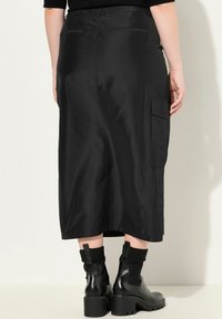 Schwarzer Midi-Rock aus satinähnlichem Material, mit glatter Textur, zwei hinteren Taschen und kombiniert mit klobigen schwarzen Stiefeln.