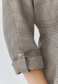 Dettaglio della manica di una camicia a quadri marrone e beige con accenti blu, caratterizzata da un tessuto testurizzato e una chiusura con bottone in legno chiaro sul polsino.