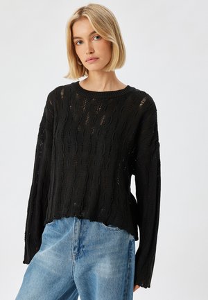 Zwarte gebreide trui met een textuurpatroon, voorzien van een ronde hals en verlaagde schouders, gecombineerd met lichtblauwe denim jeans.