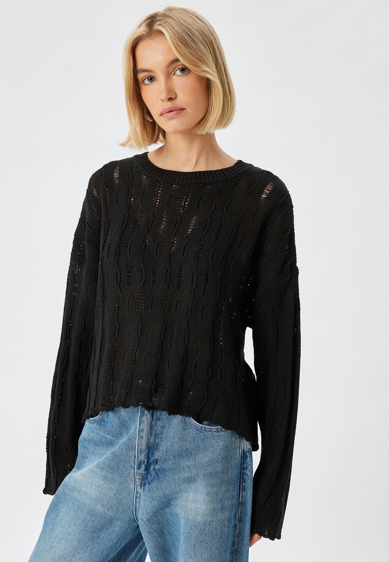 Schwarzer Strickpullover mit strukturiertem Muster, runder Halslinie und überschnittenen Schultern, kombiniert mit hellblauen Jeans.