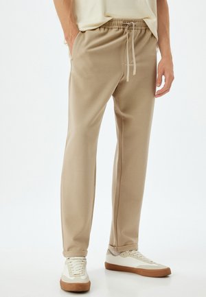 Beige sweatpants med snoretræk i taljen, sidelommer og manchetter ved anklerne, parret med hvide sneakers og en lys top. Glat tekstur.