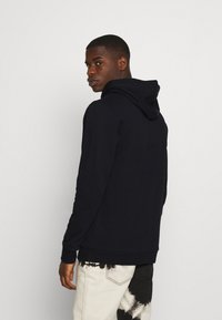 Jūrinis džemperis su raišteliu ant gobtuvo, minkšta tekstūra, ilgomis rankovėmis ir megztomis rankogaliais. Modelis dėvi šviesius tie-dye tipo kelnes.