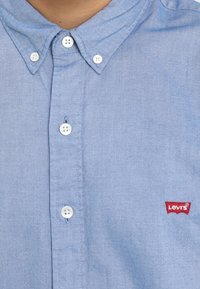 Chemise boutonnée bleu clair avec boutons blancs et une étiquette logo Levi's rouge sur le côté gauche de la poitrine.