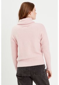 Un pull à col roulé rose clair en tissu doux, avec un col et un ourlet côtelés, porté avec un jean taille haute gris foncé, vu de dos.