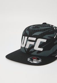 Melna beisbola cepure ar kamuflāžas rakstu zaļos toņos, ar lielu sudraba "UFC" logo priekšpusē un zīmola uzšuvi sānos.