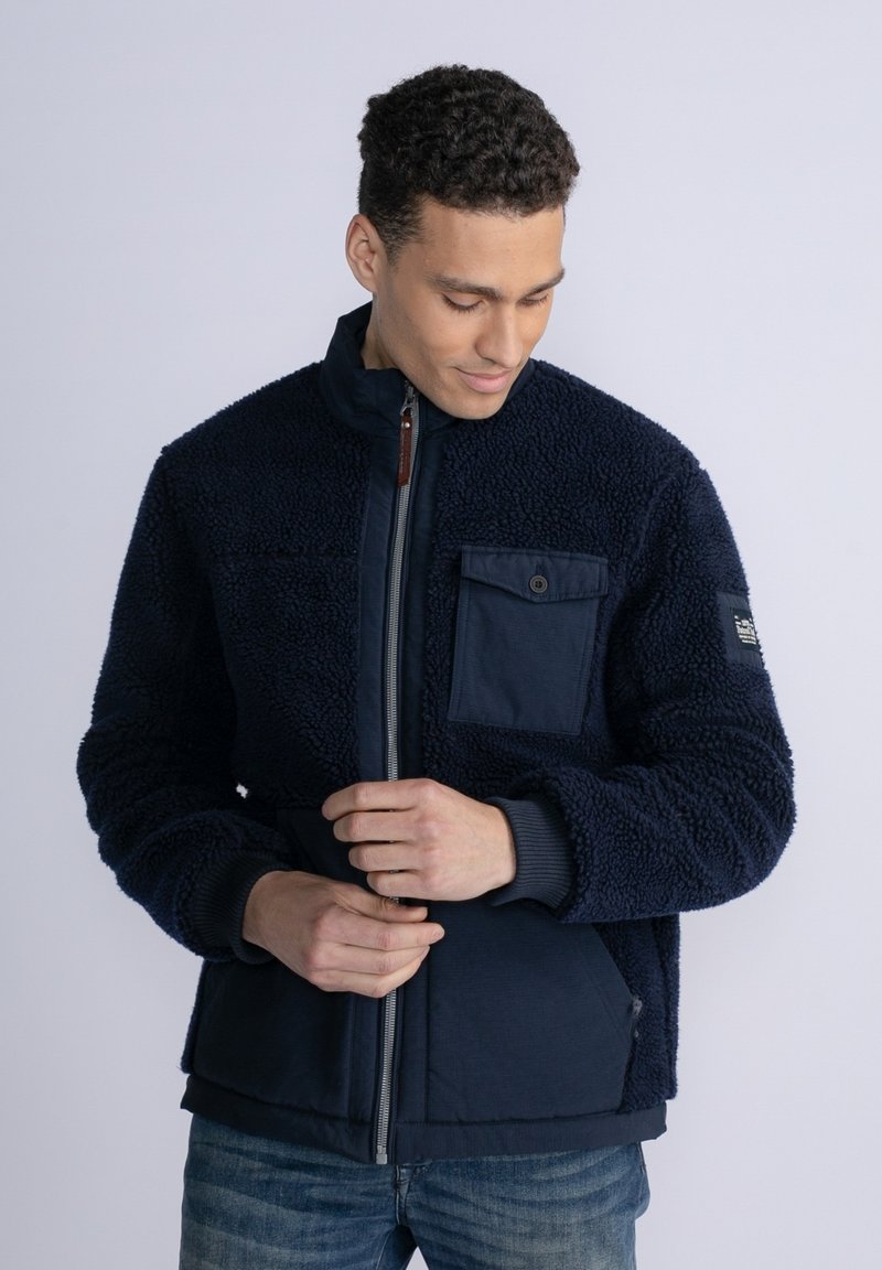 Navy fleece jacket with a textured surface, full zip, stand collar, and a chest pocket. Features ribbed cuffs and lower hem. Casual design.