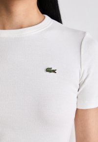 Lähedalt vaadatuna on isik, kellel on seljas valge ribaga lühikeste varrukatega särk, mille rinnal on pisike rohelise krokodilli logo tikand.