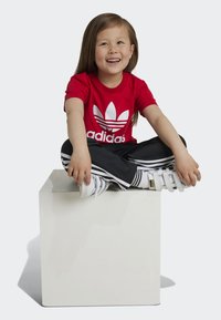 T-shirt rossa Adidas con logo bianco, pantaloni della tuta neri con strisce bianche e sneakers bianche. Seduto su un cubo bianco. Sfondo chiaro.