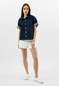 Riflová košile s vyhrnutými rukávy, přední kapsou a stříbrnými knoflíky, doplněná bílými kraťasy a bílými teniskami, na jednobarevném pozadí.