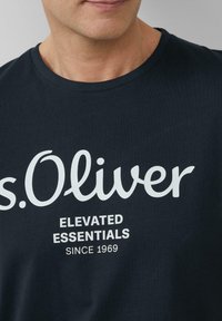 Granatowa bawełniana koszulka z okrągłym dekoltem, z białym napisem „s.Oliver” oraz wyraźnym hasłem „ELEVATED ESSENTIALS SINCE 1969”.