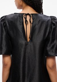 Μαύρη μπλούζα με υφή που διαθέτει φουσκωτά κοντά μανίκια και βαθύ V-λαιμό προσαρμοσμένο στην πλάτη με δέσιμο. Το ύφασμ�α έχει μια διακριτική γυαλάδα.