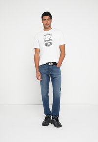 Biele grafické tričko s čiernym textom. Modré džínsové nohavice s rovný strih. Čierne tenisky s elegantným dizajnom. Jednoduchý opasok.