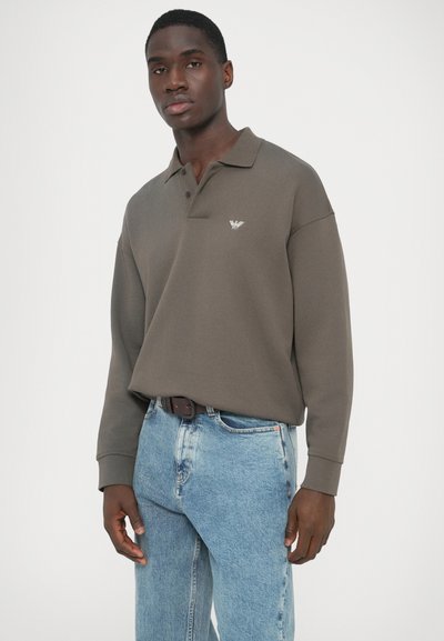 Μακρυμάνικο πόλο σε σκούρο ελαιδί πράσινο με γιακά και διακριτικό λογότυπο στο στήθος. Φοράται με ανοιχτόχρωμο τζιν παντελόνι.