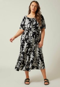 Vestido midi de estampado floral en blanco y negro con escote off-the-shoulder, cintura elástica y falda en capas. La modelo lleva sandalias negras.