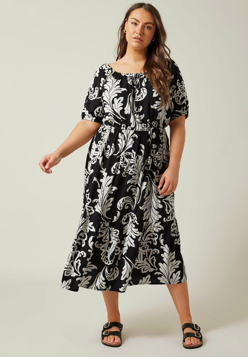 Vestido midi de estampado floral en blanco y negro con escote off-the-shoulder, cintura elástica y falda en capas. La modelo lleva sandalias negras.