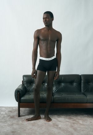 Homme debout, torse nu, portant un boxer noir Tommy Hilfiger devant un canapé en cuir vert foncé sur un sol clair tapissé.