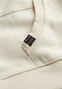 Cream-colored hoodie with ribbed cuffs. Features a black label with gold text and a logo, and visible stitching along the neckline.
