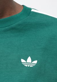 Tmavozelená mikina s okrúhlym výstrihom, s bielym vyšívaným logom troch listov a dvoma bielymi pruhmi na ramenách.