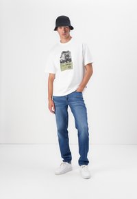 Camiseta blanca de manga corta con estampado gráfico y acentos verdes. Jeans de mezclilla azules, de corte slim. Deportivas blancas y un sombrero de cubo oscuro.