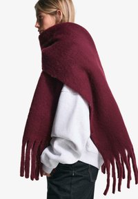 Bourgogne ullscarf med en texturerad yta, med långa fransar i ändarna, draperad över en grå sweatshirt, som visar upp en modern design.