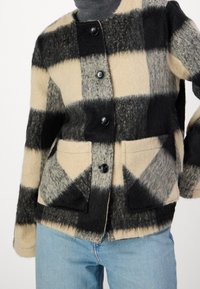 Wełniana kurtka z czarno-szaro-beżowym wzorem w kratę, okrągłym dekoltem, zapięciem na guziki oraz detalami kieszeni z przodu.
