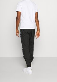 Man in zwarte sportbroek met witte gestreepte zijkanten, wit T-shirt, witte sokken en witte Adidas sneakers die wegloopt.
