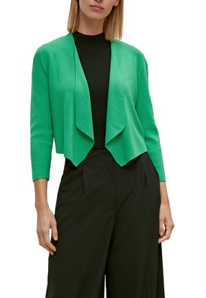 Eine Frau trägt eine leuchtend grüne offene Strickjacke über einem schwarzen gerippten Rollkragenpullover und lockeren schwarzen Hosen und steht vor einem weißen Hintergrund.