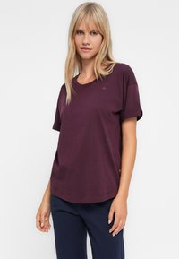 Plavolaska, oblečena v ohlapno, s kratkimi rokavi temno vijolično majico in mornarsko modre hlače, stoji ob enobarvni beli ozadju.